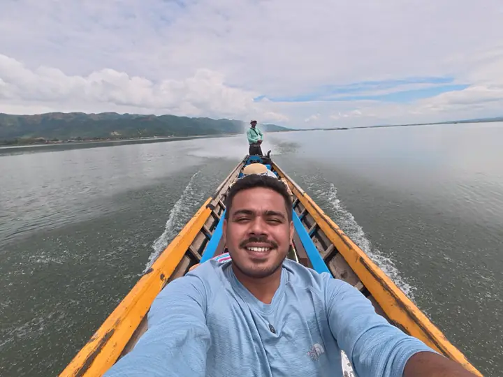 Selfie of Saif at the front of a long wooden boat speeding across Inle Lake, the boatman at the stern and hills on the horizon