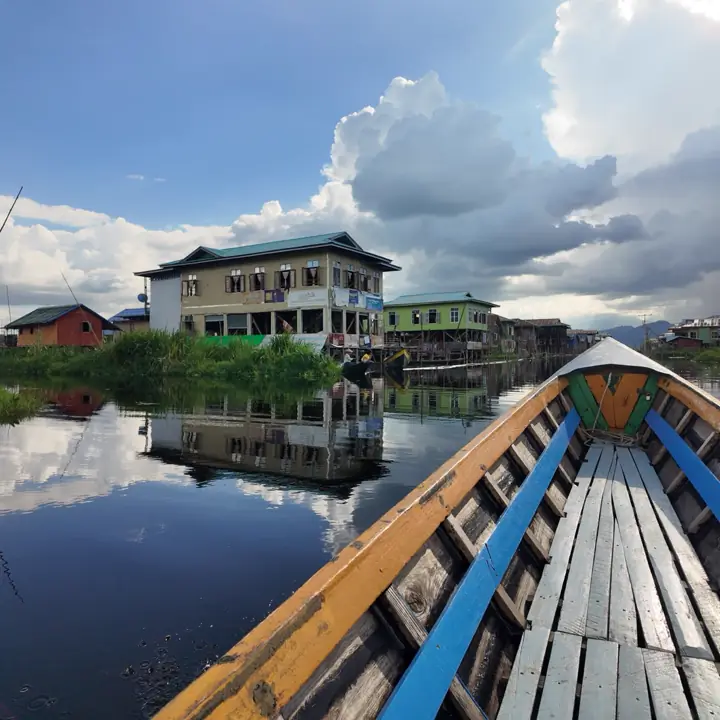 Bow of a wooden longtail boat heading toward stilt houses reflected in the still water of Inle Lake
