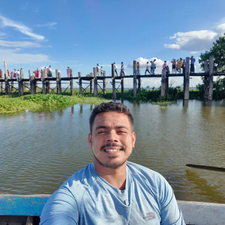Saif in a boat below U Bein Bridge, the long teak footbridge crowded with people against a blue sky over Taungthaman Lake